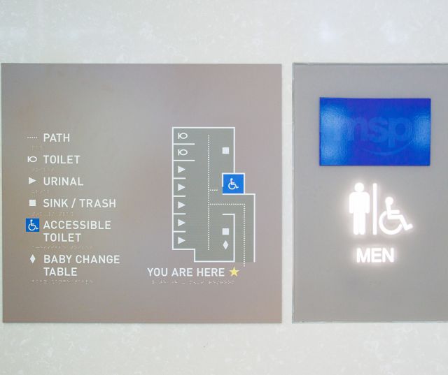 A tactile wayfinding map with raised symbols and braille labels for restroom features, next to the men's restroom at MSP airport.